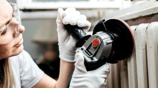Woman wearing safety goggles and white gloves using an angle grinder to sand a metal column radiator