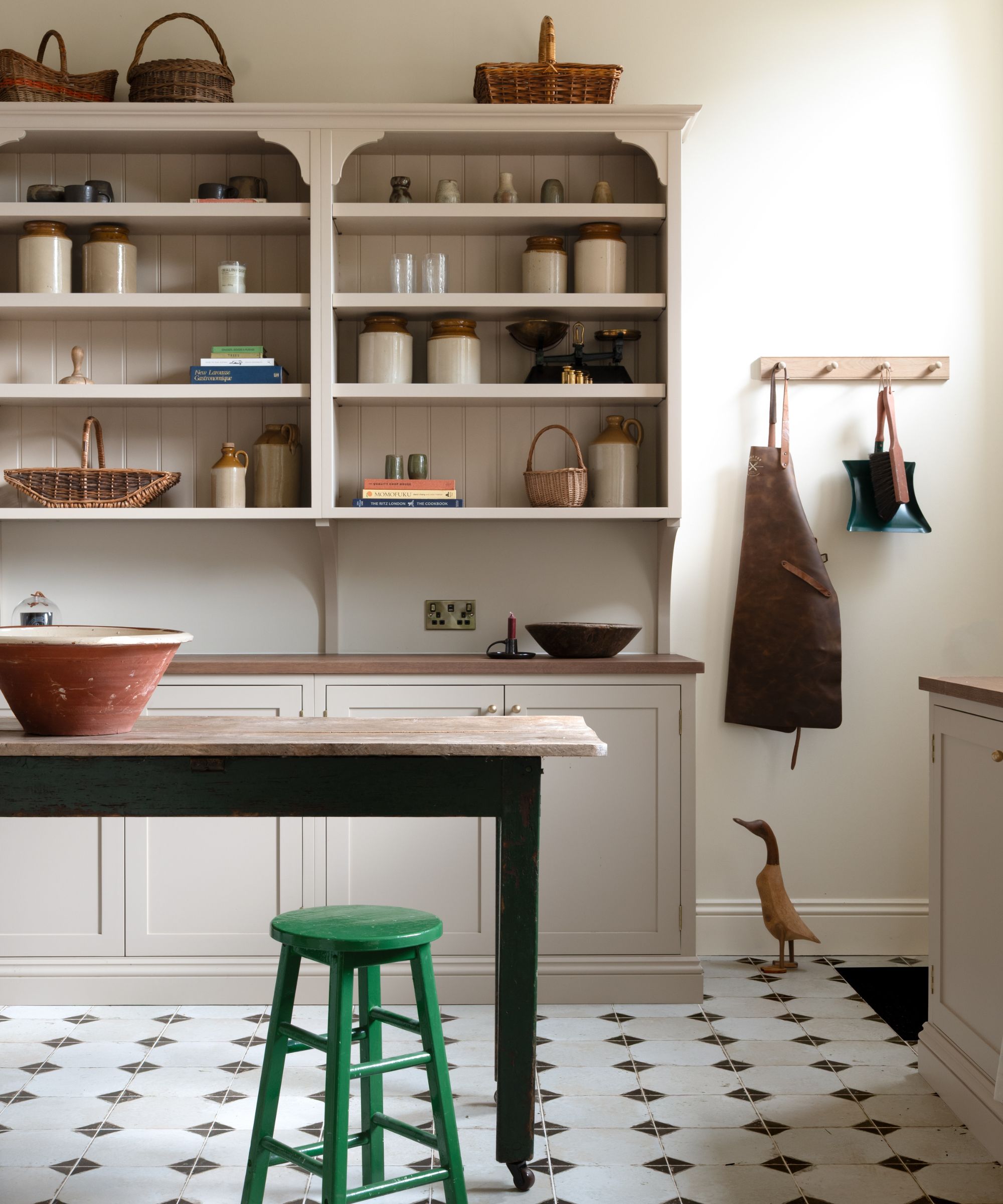 Shaker-style pantry with tiled floor, a large dresser with shelves of ceramics and kitchen utensils, a wooden table with a green stool beneath it, and pegs on the wall holding an apron.
