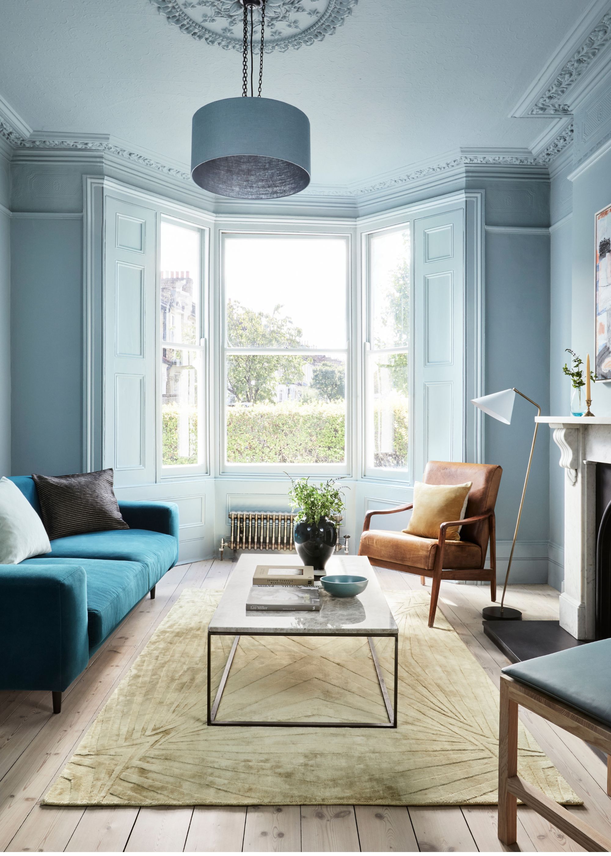 A living room painted light blue with a large bay window and a dark blue sofa, a brown armchair, and a light green rug with a modern coffee table.