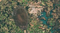 A satellite photo of a giant dark dome next to a town and an artificial lake, all surrounded by patchwork fields