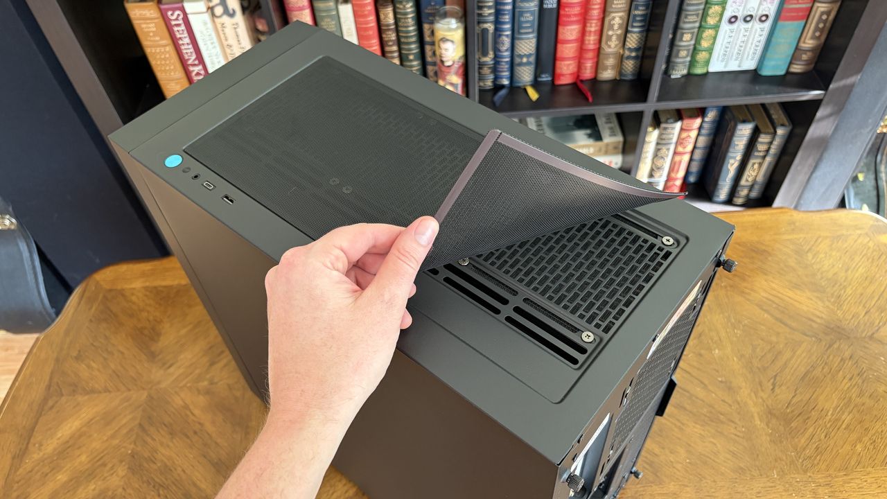 A hand lifts a mesh dust filter from the top of a sleek Maingear MG-1 MK.II computer tower on a wooden surface, with a bookshelf filled with colorful books in the background.