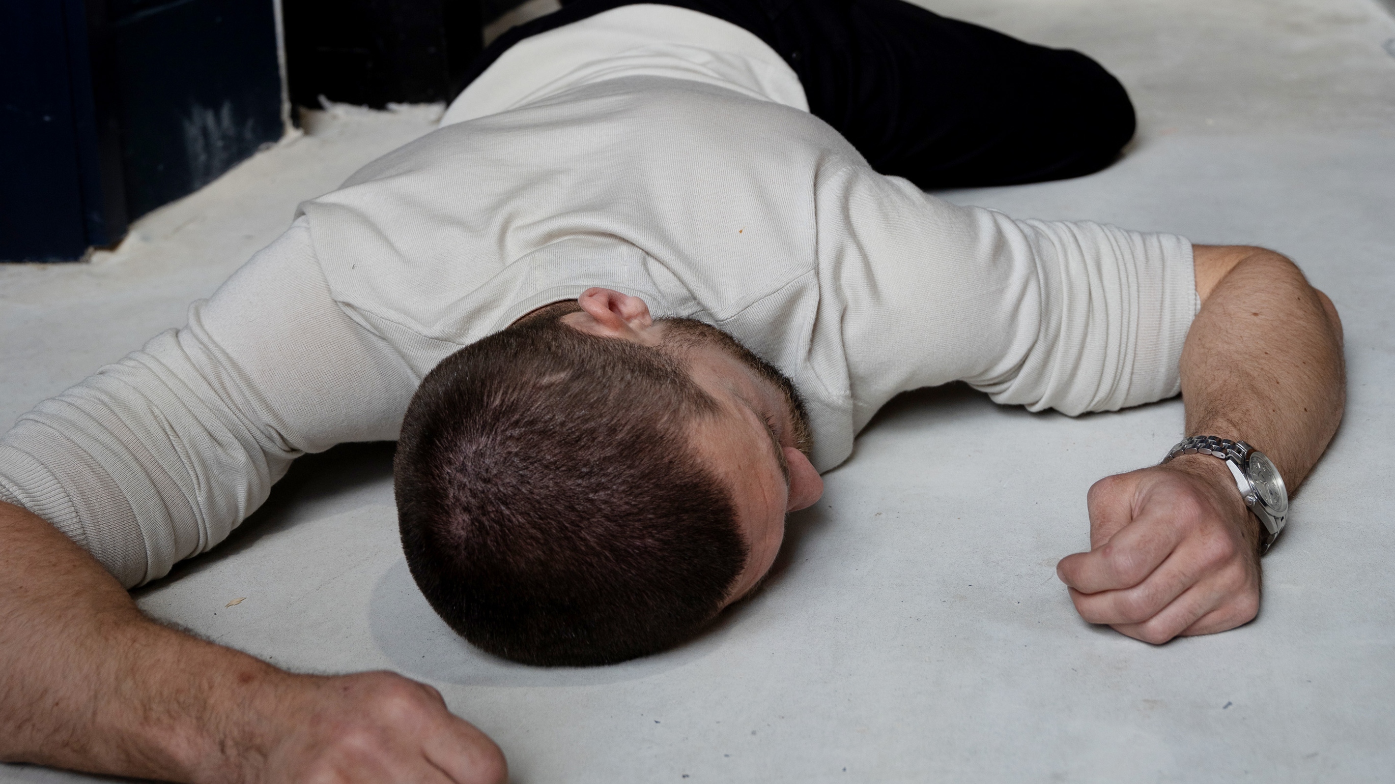 Joe Take lying on his front on the floor, unconscious, and wearing a white tshirt 
