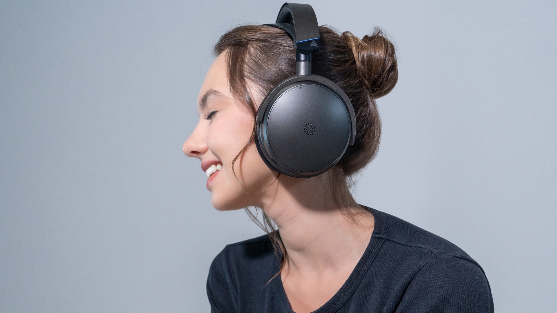 A profile view of a woman smiling while wearing large, dark gray over-ear headphones with a blue accent on the headband.