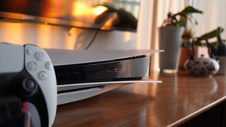En PlayStation 5 og en DualSense-kontroller står ved siden av hverandre på et TV-stativ på en solrik dag.