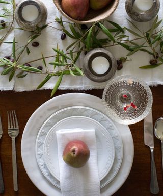 A Christmas table setting with white plates, a linen table runner topped with mistletoe and candles, and silver cutlery