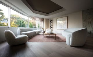 The lounge featuring two white curved extended sofas and a chair, a brown rug, a double layer table, large panoramic windows that look out to trees.