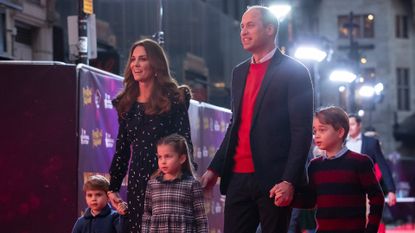 Kate Middleton wears a long-sleeved polka dot dress while Prince William wears a suit jacket with a red sweater, as they hold hands with their kids, Prince George, Princess Charlotte, and Prince Louis