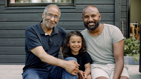 A smiling grandfather sits on a deck with his son and granddaughter.