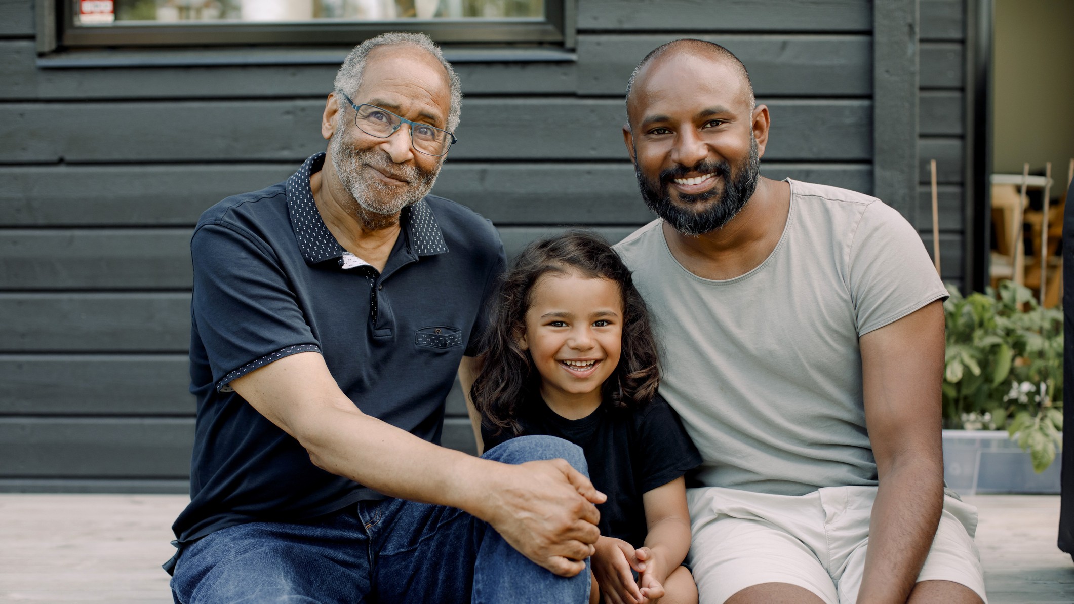 A smiling grandfather sits on a deck with his son and granddaughter.