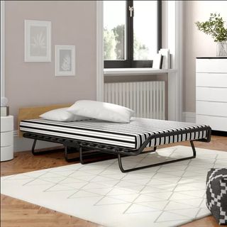 a basic folding bed with wooden headboard, a black frame, and a black and white mattress, in a room with a light wooden floor, a white rug, pink walls, a white chest of drawers, and a window with black frames