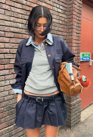 A woman wearing a blue jacket over a blue button-down shirt and gray polo styled with a navy pleated miniskirt and brown bag.