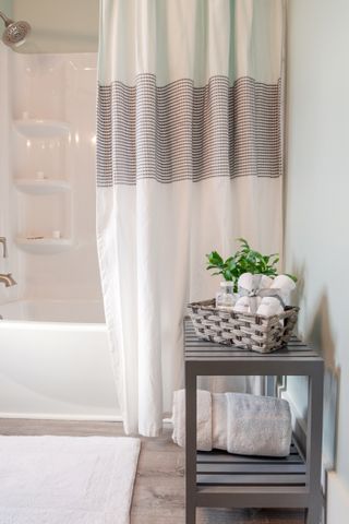 A modern white bathroom with a white shower curtain with grey stripes on, next to a small grey side table with a basket on top.