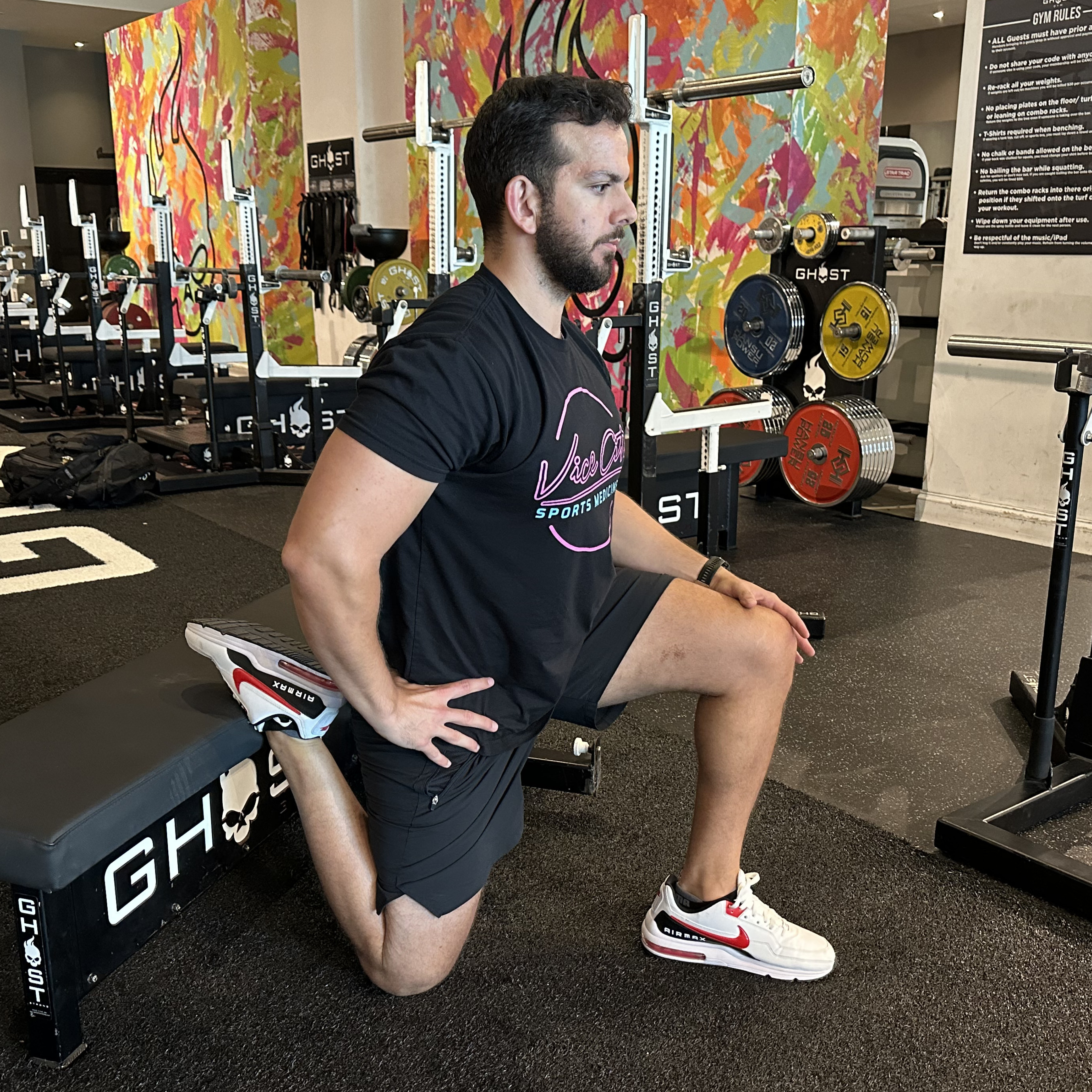 An employee at Vice City Sports Medicine demonstrates a couch stretch in a gym setting. They are directly in front of a weights bench, with their right shin resting on the bench, knee on the floor, left foot forward in a half-kneeling stance. Their torso is upright.