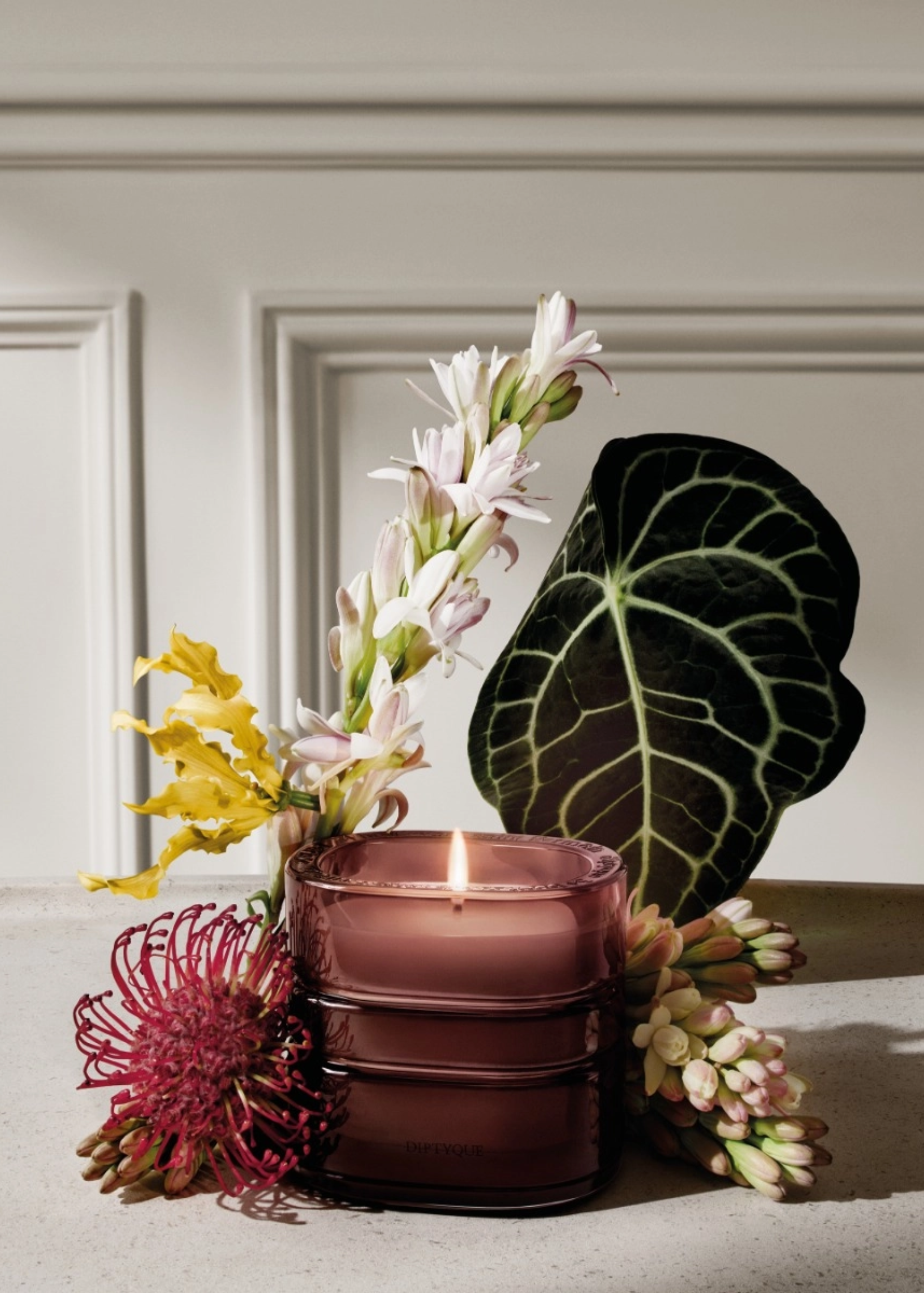 A brown glass Diptyque candle with leaves and colorful flowers surrounding it