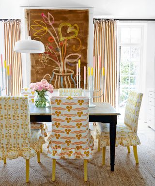 Yellow dining room with contrasting prints and patterns, all united through the use of yellow. White painted walls, yellow striped curtains, large hanging mural, painted design with flowers, dark wood rectangular dining table with chairs, upholstered with yellow and white printed designs, yellow painted feet, textured natural carpet, low hanging white pendant over table, table decorated with large candleholder and pink flowers