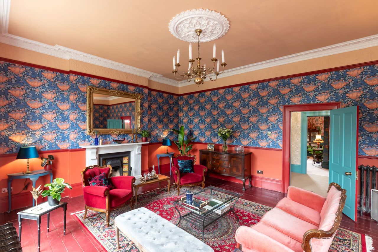 The period style living room has claret coloured sofas and green patterned wallpaper whilst a black fireplace sits by the side