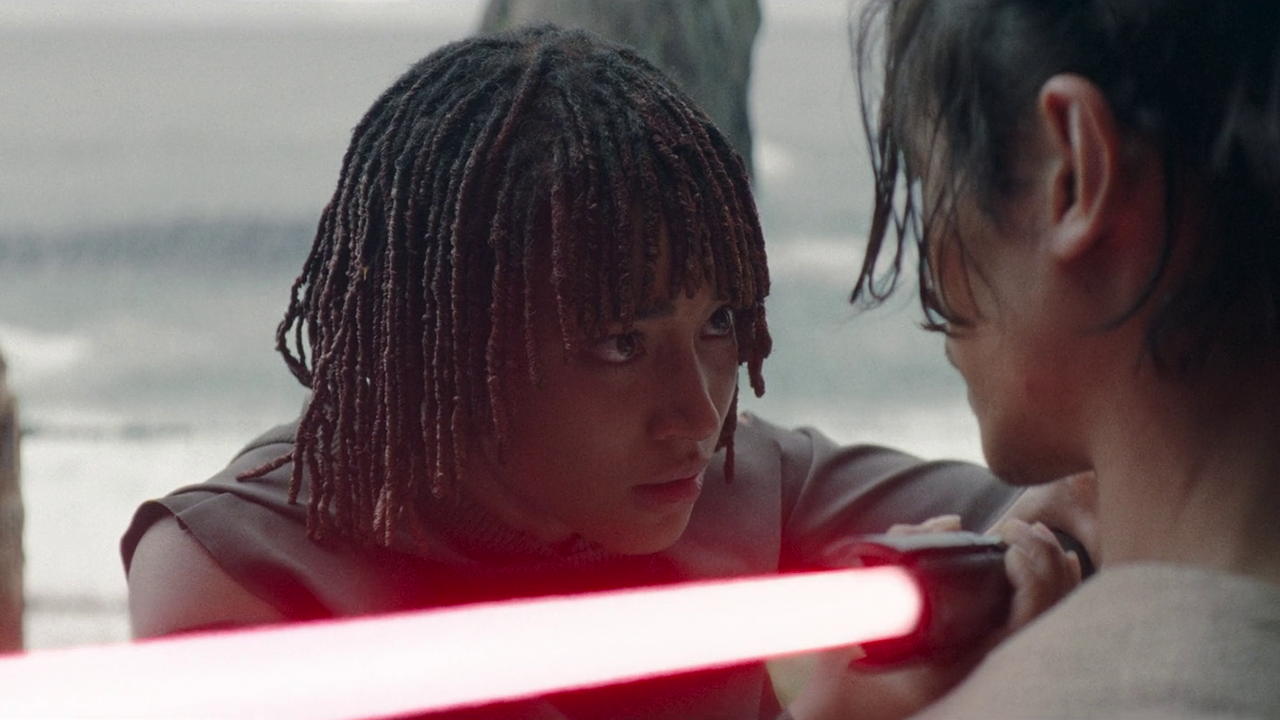 A young woman holds a red lightsaber threateningly against the neck of another person.