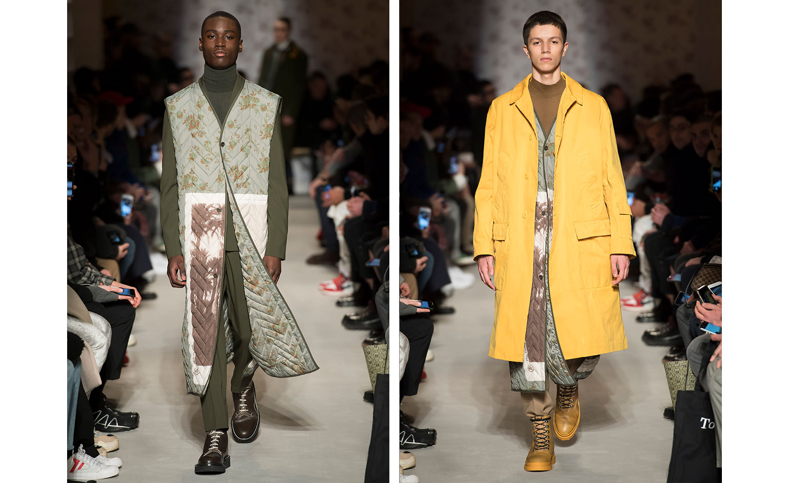 Catwalk view of two models wearing looks from OAMC's collection. One model is wearing a dark green high neck top, dark green suit, a long light green and white piece with floral pattern and brown boots. And the second model is wearing a brown top, long light green and white piece with floral pattern, yellow coat and dark yellow boots. There are people seated on both sides of the catwalk