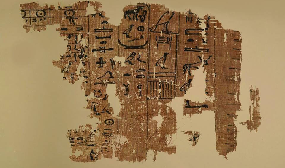 A rectangular but fragmented piece of brown papyrus with hieroglyphs written on it in black ink.