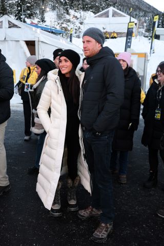 Meghan Markle wearing a long white coat and snow boots standing next to Prince Harry wearing a black coat and gray hat in front of white tents and a snowy mountain