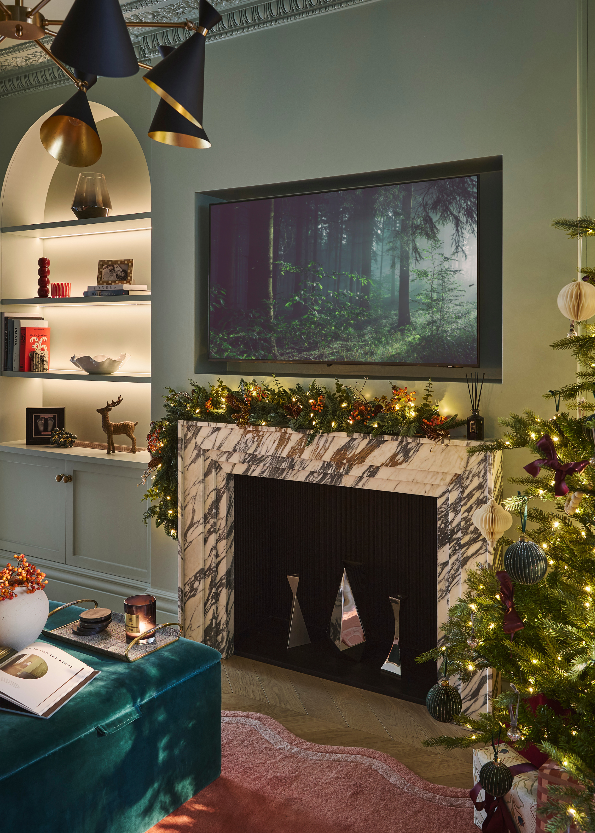 A modern living space decorated for christmas with a forest scene on the tv above the decorated mantelpiece with a lit garland with a lit and decorated christmas tree partly in view