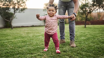 A toddler takes her first steps while outside on the lawn with her mother right behind her.