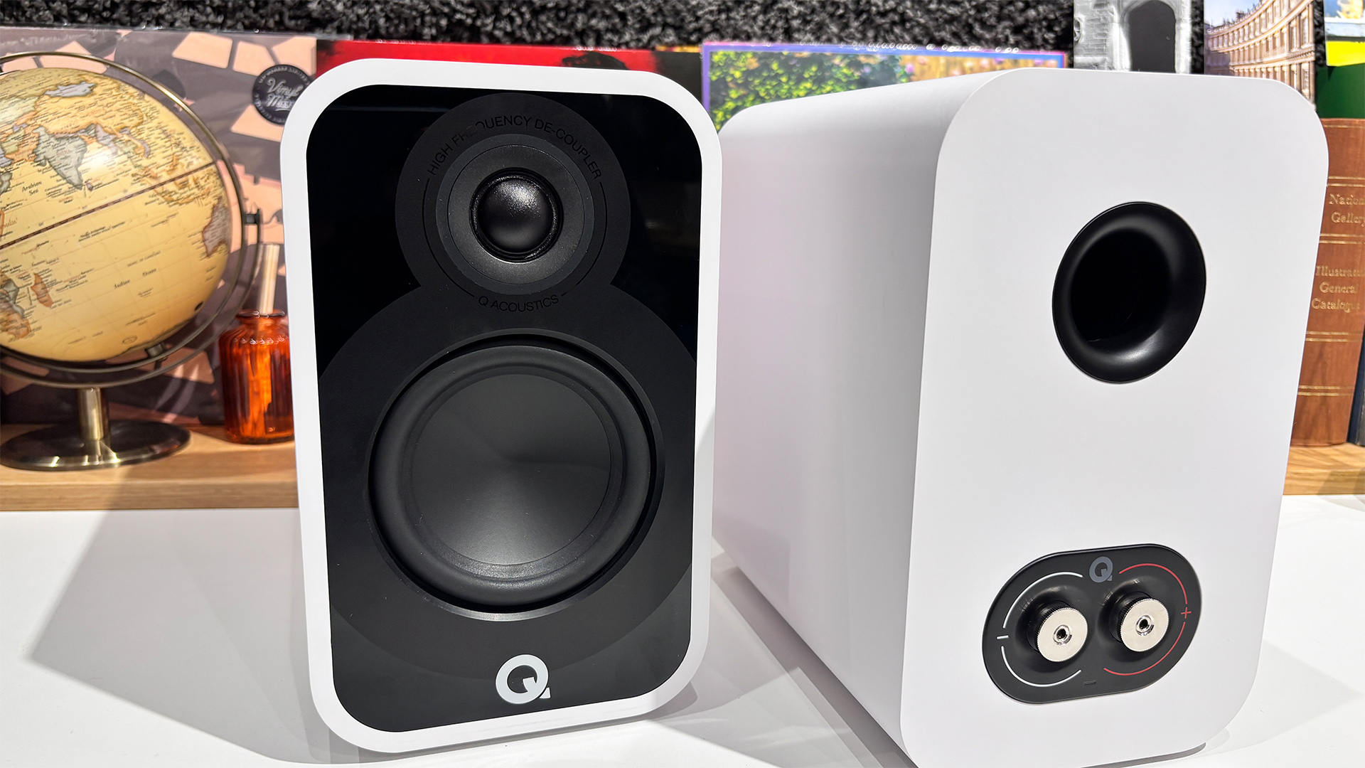 Q Acoustics 5010 standmount speakers on white surface in front of busy shelf, one of the speakers is turned around