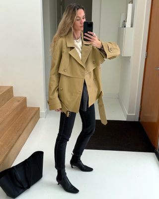 influencer Anouk Yve poses for a mirror selfie wearing a cropped trench coat, white top, skinny black jeans, and black square-toe kitten heel boots