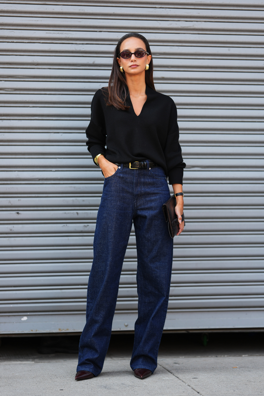 A guest wears straight dark brown hair parted in the center with a black fabric headband, small black oval sunglasses, chunky gold hoop earrings, a black leather strap watch with a gold case on the wrist, a black croc-embossed leather envelope clutch, a black long-sleeve V-neck knit wool sweater slightly tucked at the front, a black leather belt with a gold rectangular buckle fastening high-waisted dark navy blue wide-leg denim trousers, and black pointed-toe leather pumps with a low heel, outside COS, during New York Fashion Week