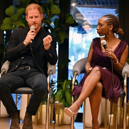 Prince Harry speaking into a mic sitting on stage next to Sophie Chandauka