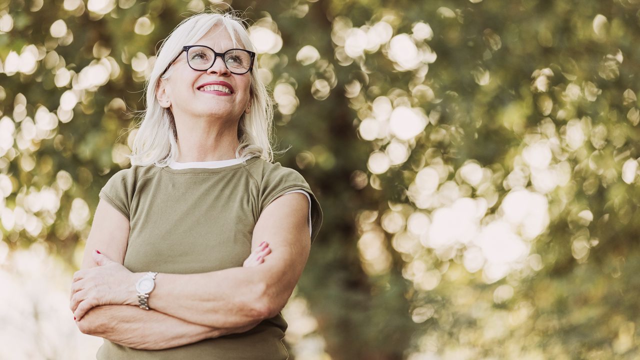 An confident-looking older woman stands outside under a tree with her arms crossed.