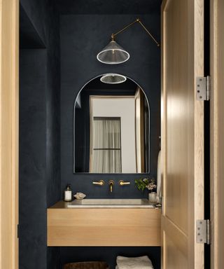 powder room with dark navy walls, wood door and sink, arched mirror with modern glass wall sconce