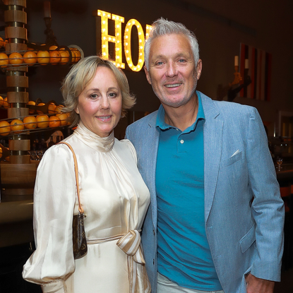 Shirlie on the left wearing white silk dress and Martin is on the right in blue polo shirt and blazer. They have an arm around each other.
