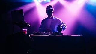 Man DJs on a dark stage with a pink and purple light behind him