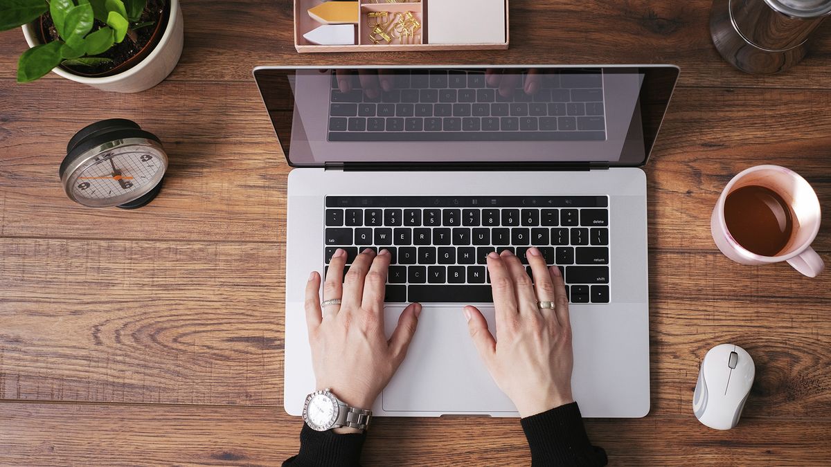 Person typing on a laptop on a desk with cup of tea next to it.