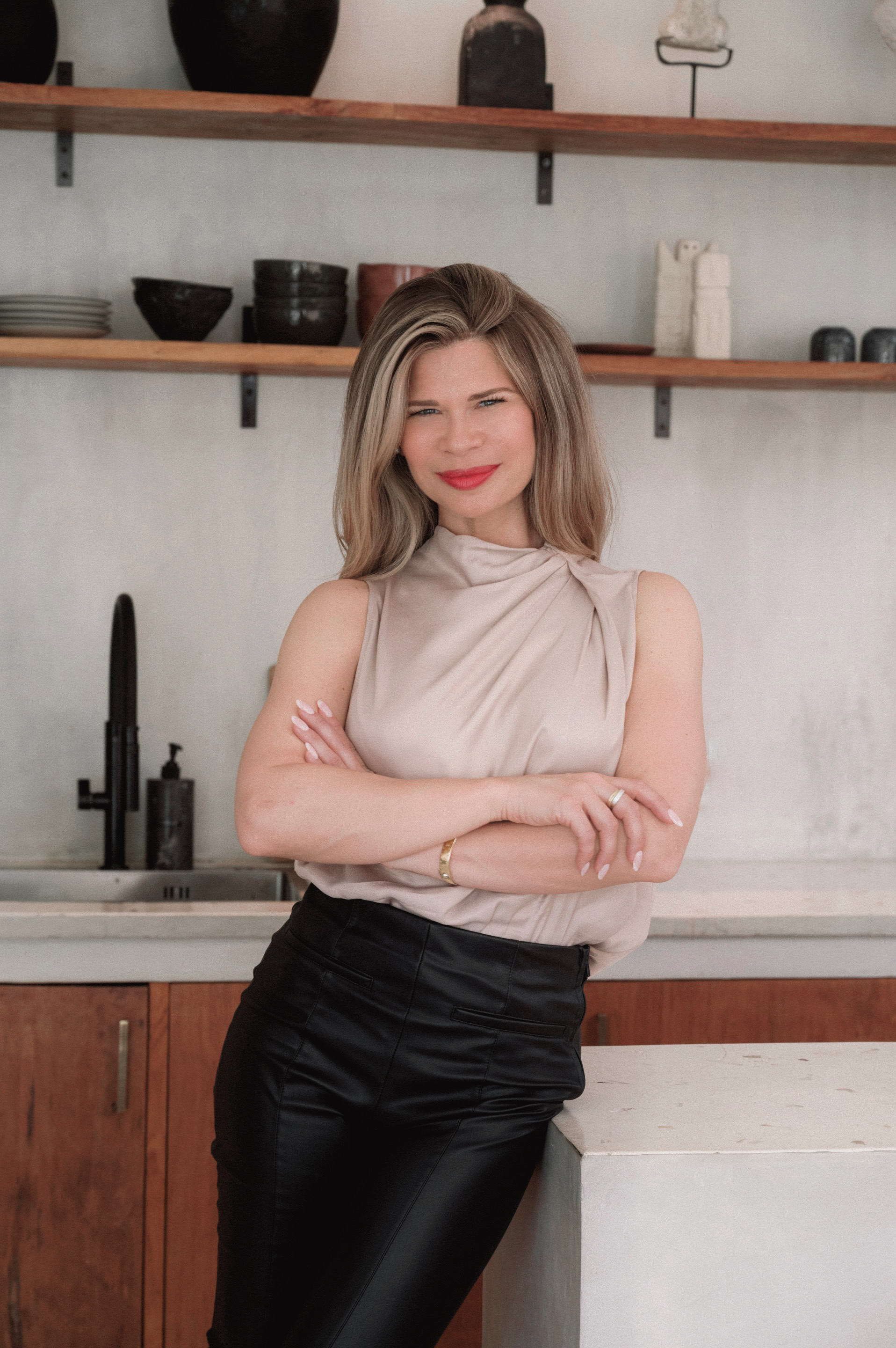 a headshot of a white woman with bronde hair wearing a beige blouse and black pants. she is standing with her arms crossed