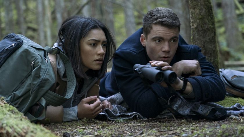 Rose and Peter crouched down in forest, sharing a pair of binoculars, in The Night Agent