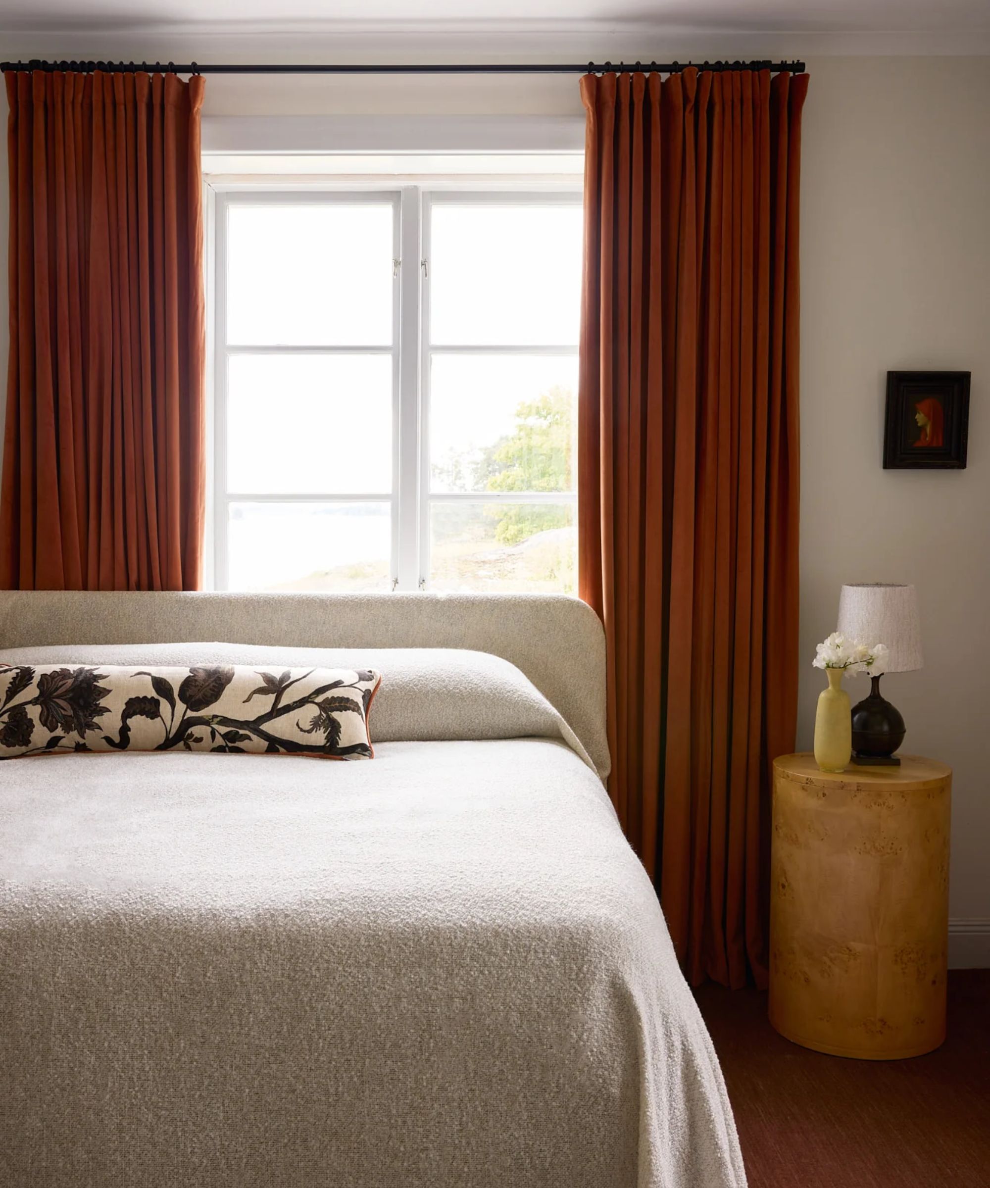 neutral bedroom with velvet terracotta curtains and a linen bed in front of the mirror, with a burl wood circular shaped bedside table with small lamp