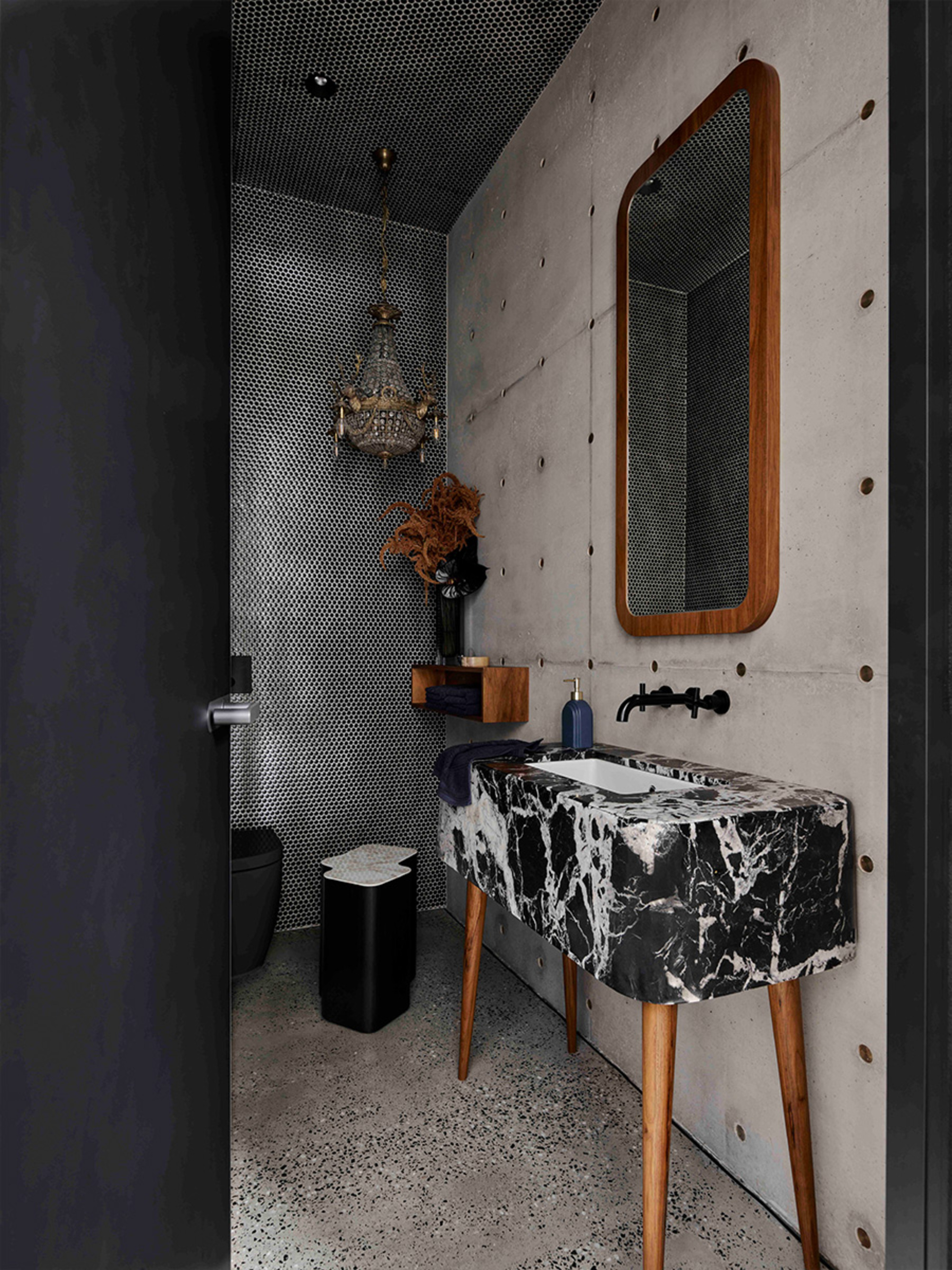 mid century modern bathroom with off-form concrete, a freestanding black marble vanity, timber mirror, black tapware, mosaic walls, chandelier, and black door