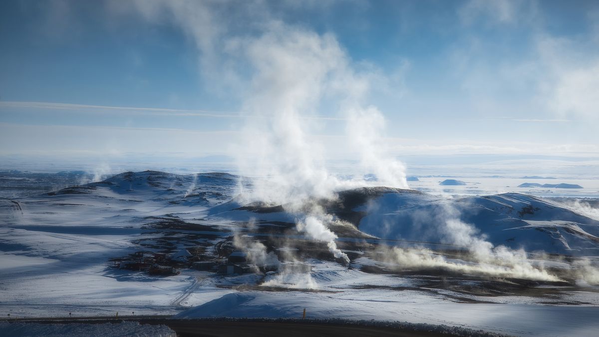 An aerial shot of the Krafla volcanic caldera in Iceland, which is used for geothermal green energy. Steam rises from the fissure in the earth, with pale blue water contrasted against the dark rocks.