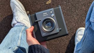 Fujifilm Instax Wide Evo camera held in a hand with a persons legs and shoes in the background