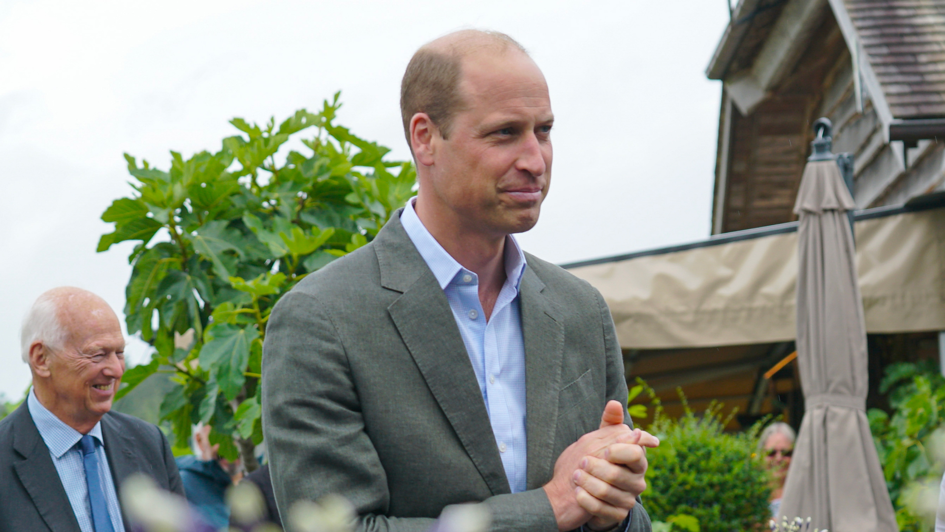 Prince William wears a gray suit and light blue tie and holds his hands together while looking pensive