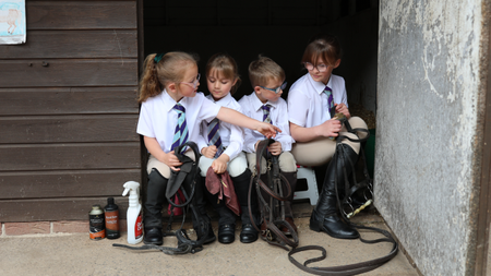 Four children sit in a ack room doorway cleaning tack at Pony Club camp. They wear riding boots, beige jodhpurs, white shirts and blue and purple ties