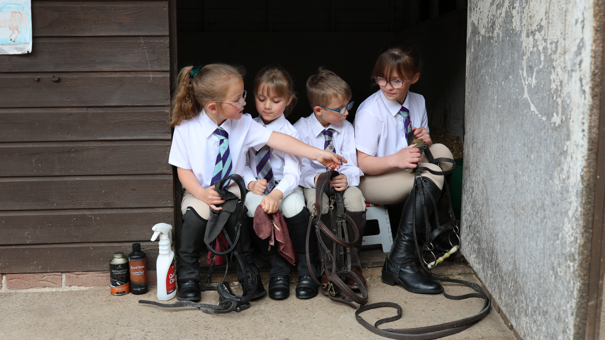 Four children sit in a ack room doorway cleaning tack at Pony Club camp. They wear riding boots, beige jodhpurs, white shirts and blue and purple ties