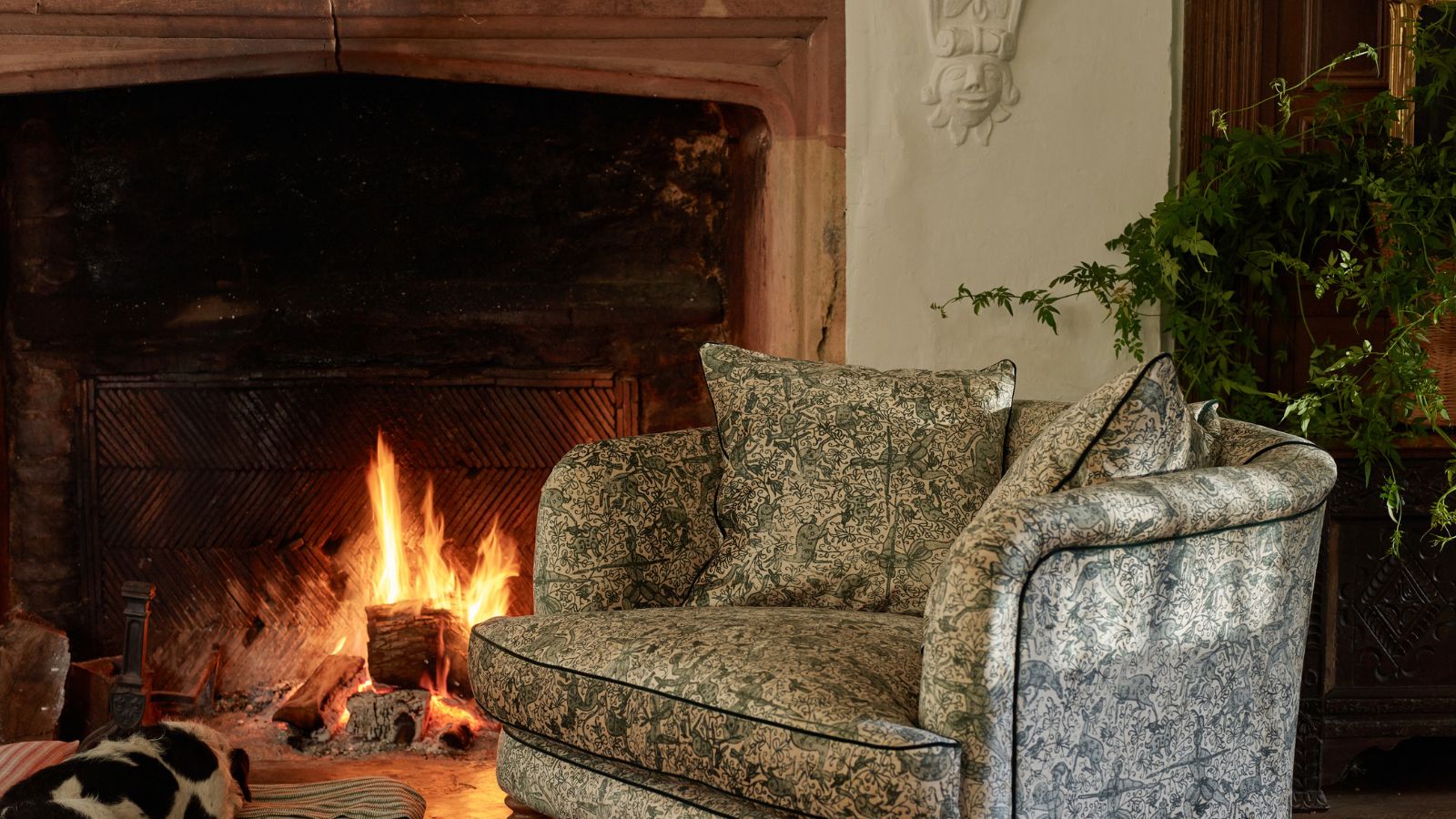 A paisley print upholstered tub armchair by a cozy fireplace with a stone mantel surround. There is a black and white dog asleep on a fabric bed by the fire