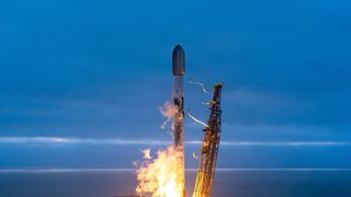 A white and black rocket launches into an overcast evening sky from an ocean-side pad.
