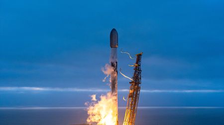 A white and black rocket launches into an overcast evening sky from an ocean-side pad.