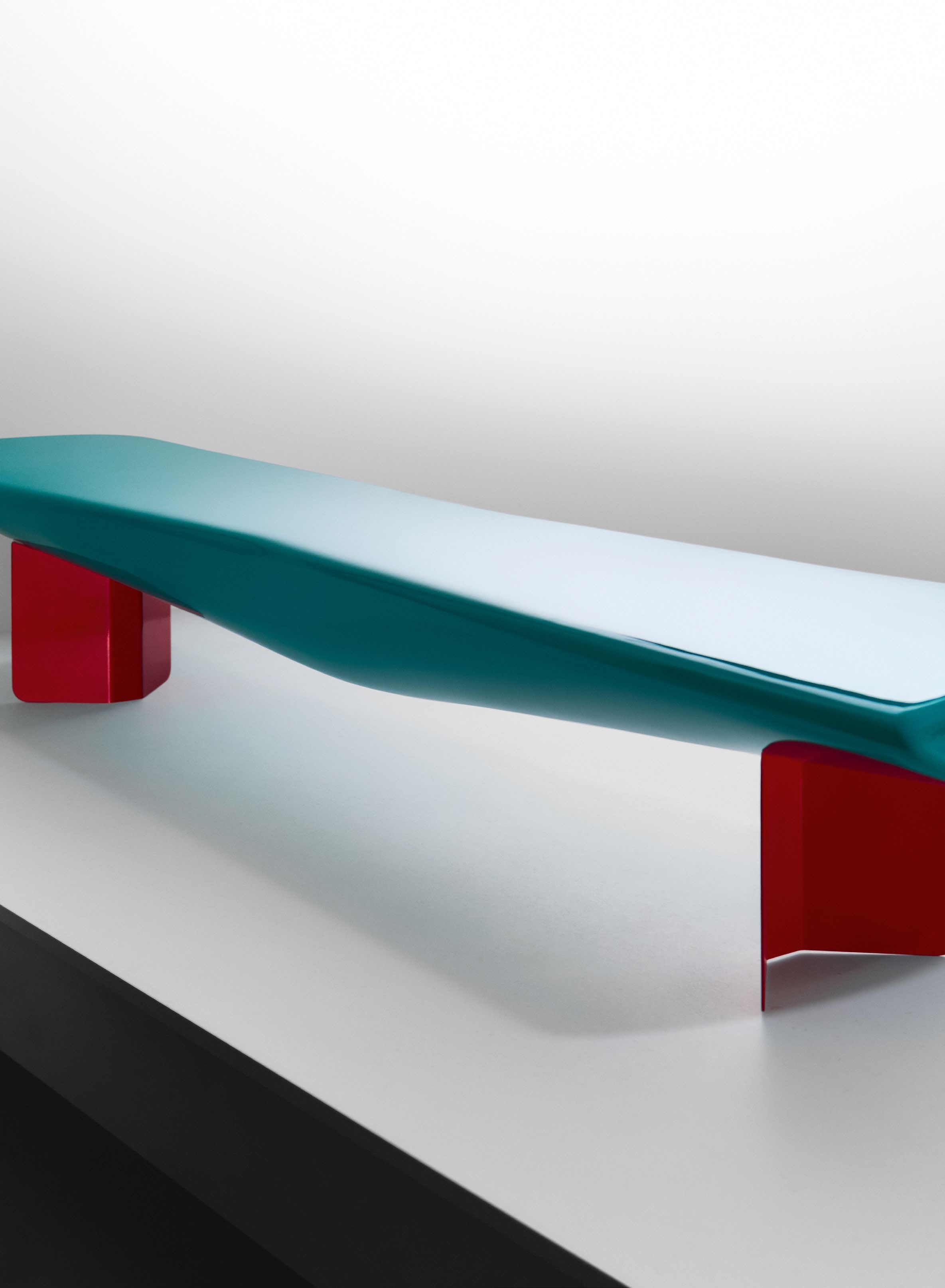 A contemporary bench designed by Ajay Shah, featuring a smooth turquoise fibreglass top supported by bold red metal legs, showcased in a minimalist setting.