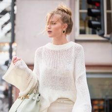 A young woman crosses the street wearing a light cream knitted sweater and carrying a mint green bag.