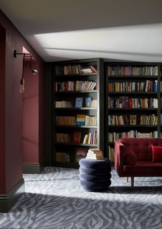 basement living room with grey zebra style carpet, round grey footstool, claret red velvet sofa, bookcases, claret red walls, white ceiling, wall lights